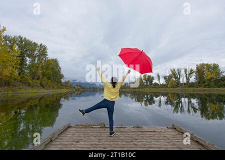 Una donna che tiene un ombrello rosso si mette in posa mentre si trova su un molo vicino al fiume Kootenai nell'Idaho settentrionale. Foto Stock