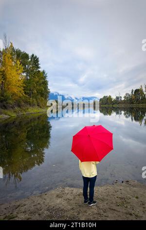 Una donna con un ombrello rosso si trova sulle rive del tranquillo fiume Kootenai vicino a Bonners Ferry, Idaho. Foto Stock
