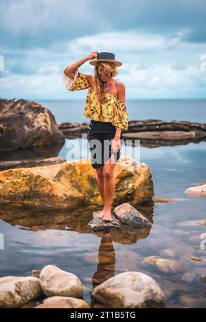 Ragazza bionda caucasica in camicia floreale, pantaloncini neri e cappello di paglia in un paesaggio naturale vicino al mare e alle rocce al tramonto, stile di vita. Nascosto nel Foto Stock