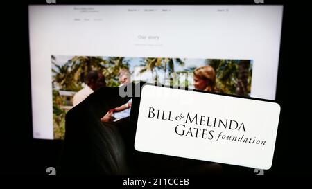 Persona in possesso di un cellulare con il logo dell'American Bill and Melinda Gates Foundation (BMGF) davanti alla pagina web. Concentrarsi sul display del telefono. Foto Stock