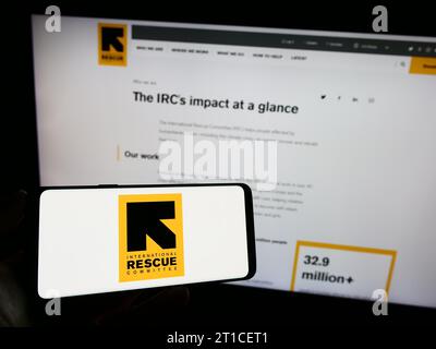 Persona in possesso di un cellulare con il logo dell'organizzazione International Rescue Committee (IRC) davanti alla pagina web. Concentrarsi sul display del telefono. Foto Stock