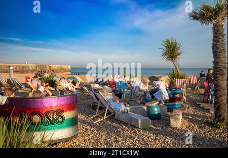 Le persone si siedono su sdraio in una giornata di sole sulla spiaggia al Pilot Beach Bar di Folkestone, Kent, Regno Unito. Foto Stock