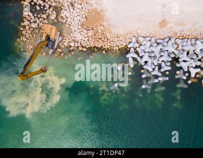 Un escavatore costruisce diligentemente un molo o un frangiflutti nel mare, il suo potente braccio che si estende dalla riva, creando una struttura resiliente in mezzo Foto Stock