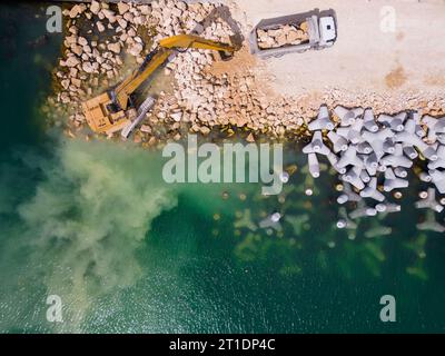 Un escavatore costruisce diligentemente un molo o un frangiflutti nel mare, il suo potente braccio che si estende dalla riva, creando una struttura resiliente in mezzo Foto Stock