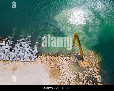 Un escavatore costruisce diligentemente un molo o un frangiflutti nel mare, il suo potente braccio che si estende dalla riva, creando una struttura resiliente in mezzo Foto Stock