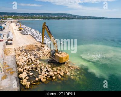 Un escavatore costruisce diligentemente un molo o un frangiflutti nel mare, il suo potente braccio che si estende dalla riva, creando una struttura resiliente in mezzo Foto Stock