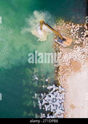 Un escavatore costruisce diligentemente un molo o un frangiflutti nel mare, il suo potente braccio che si estende dalla riva, creando una struttura resiliente in mezzo Foto Stock