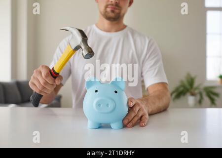 Il giovane tiene il martello e vuole rompere una scatola di denaro a forma di maialino blu con i suoi risparmi Foto Stock