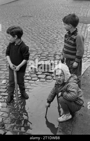 Bambini che giocano con i bastoni in una pozzanghera sotto la pioggia su una strada acciottolata durante la slum clearance e la demolizione di St Ann's, Nottingham. !969-1972 Foto Stock