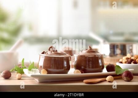 Due vasetti di vetro pieni di crema di cacao con nocciole sul piatto con foglie e nocciole intorno al banco da cucina in legno. Vista frontale. Foto Stock