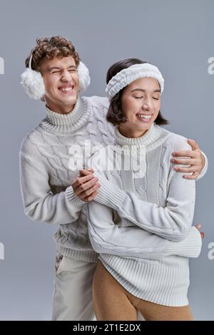 uomo felicissimo con comode cuffie che abbracciano la donna asiatica in maglione invernale grigio, stile stagionale Foto Stock