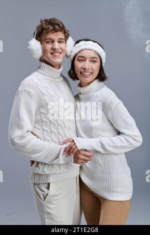 coppia interrazziale con comodi maglioni in maglia che si stagliano sullo sfondo grigio con la neve e un inverno alla moda Foto Stock
