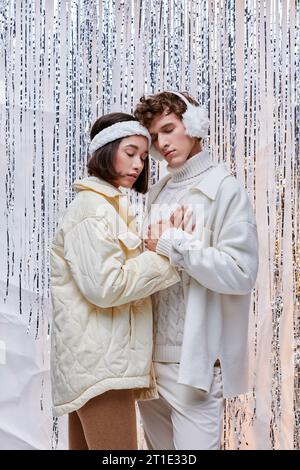 spirito natalizio, coppia interrazziale alla moda che abbraccia con occhi chiusi sullo sfondo lucente di tinsel Foto Stock