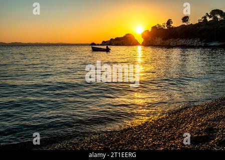 Tramonto sul mare. Il sole tramonta tra due rocce all'orizzonte, con una piccola barca a motore ancorata nelle vicinanze Foto Stock