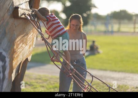 Un simpatico biondo che sale la scala di corda sul parco giochi e sua madre lo sta osservando. Scatto medio Foto Stock