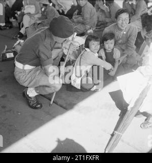 La rioccupazione britannica di Hong Kong Regimental Sergente Maggiore Redman di Aclesey nel Bedfordshire, colloqui con due bambini giapponesi che sono tra un gruppo di giapponese internati civili in attesa allo scalo per trasporto a motore che li porterà a Stanley Camp, Hong Kong. Circa 3 mila civili giapponesi sono stati spostati da Kowloon a Stanley Camp, che era stato utilizzato durante la guerra dalle truppe giapponese come un campo di internamento per Allied civili. Foto Stock