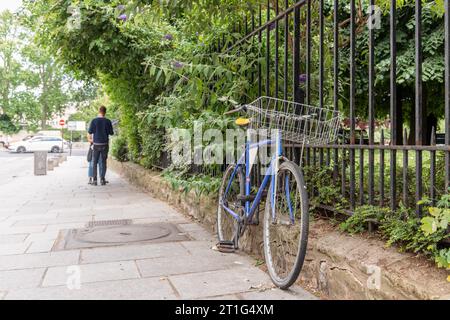 La bicicletta blu è incatenata alle ringhiere a Parigi, in Francia. Sullo sfondo sfocato, due persone sono in conversazione. Macchine parcheggiate in lontananza. Foto Stock