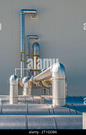 Tubazioni metalliche isolate riflettenti sul tetto di un impianto di stoccaggio a freddo (refrigerazione industriale), con tubazioni che attraversano una parete. Foto Stock