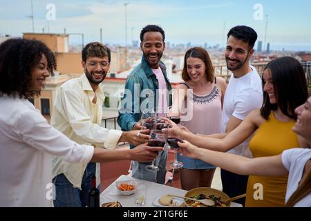 Sorridenti giovani multietnici che brindano bicchieri di vino in piedi per festeggiare i compleanni sul tetto. Foto Stock