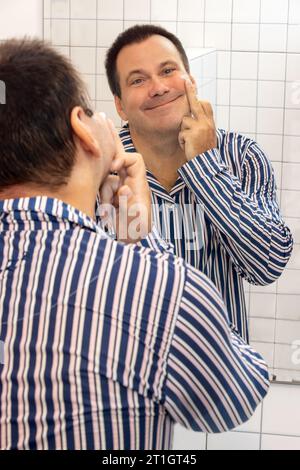 Un uomo in pigiama creme la faccia davanti a uno specchio del bagno Foto Stock