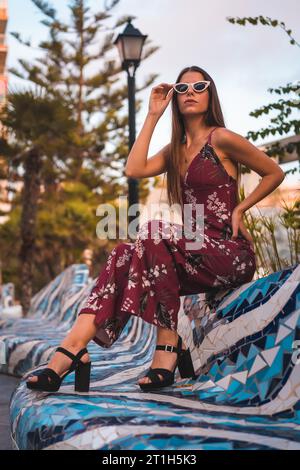 Street Style, una giovane bruna in abito floreale marrone e moderni occhiali da sole bianchi che si godono l'estate durante l'ora d'oro in città Foto Stock