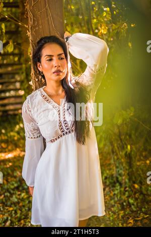 Stile di vita, bruna con capelli lisci in un bellissimo vestito bianco in un parco che riposa in estate. Ritratto appoggiato su un legno Foto Stock