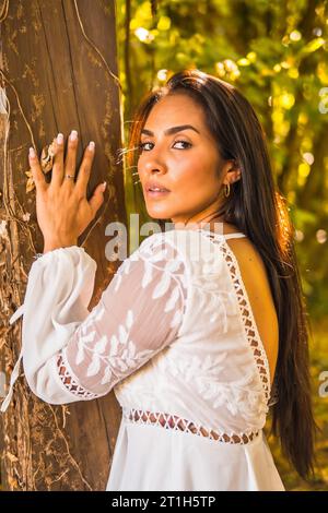 Stile di vita, bruna latina dai capelli lisci con un bellissimo vestito bianco in un parco naturale. Ritratto senxuale appoggiato su un albero molto bello Foto Stock