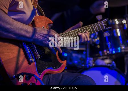 Vista ravvicinata delle mani di un uomo anziano che suona la chitarra in una jam blues in un pub di Londra con la batteria sullo sfondo Foto Stock