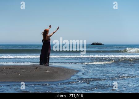 Vista posteriore di Happy Redhead Woman in lungo abito viola scuro in piedi sulla spiaggia di sabbia estiva con le braccia alzate che si estendono verso il cielo blu e le onde del mare. Grata Foto Stock