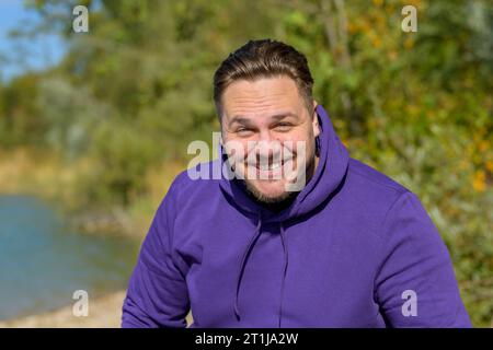 Ritratto di un giovane uomo attraente che indossa una felpa viola con cappuccio che ride divertente guardando la fotocamera, all'aperto in autunno Foto Stock