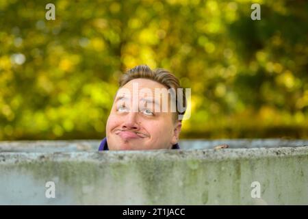 Primo piano di un giovane in piedi dietro un muro di cemento rotondo con solo la testa visibile e facendo un volto, fuori in autunno Foto Stock