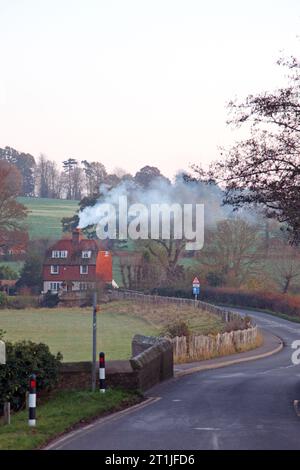 Una casa nella campagna britannica riscaldata dal legno bruciato mentre il fumo esce dal camino inquinando l'ambiente in un momento di cambiamento climatico, gas Foto Stock