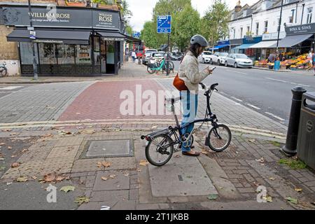 Giovane ciclista che indossa un casco da ciclismo che invia messaggi sul cellulare in piedi con la bici Brompton su Lordship Lane a East Dulwich South London SE22 Foto Stock