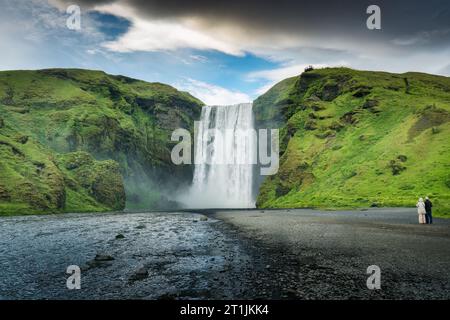 Maestosa e potente cascata Skogafoss che scorre con coppie di turisti che si divertono in estate in Islanda Foto Stock