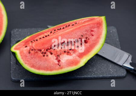 Un ritratto di un quarto di taglio di un cocomero verde adagiato su un tagliere di plastica accanto a un coltello. La fetta o il pezzo di frutta è pronto per essere mangiato Foto Stock