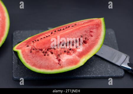 Un ritratto di un quarto di taglio di un cocomero verde adagiato su un tagliere di plastica accanto a un coltello. Il pezzo o la fetta della frutta è pronta per essere mangiata Foto Stock