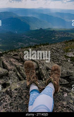 Sosta durante un'escursione in montagna. I piedi del viaggiatore in vecchi scarponi da trekking marroni su uno sfondo roccioso e boscoso della montagna. Foto Stock