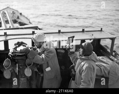 Gli ufficiali sul ponte della HMS LEGION durante il raid su isole Lofoten in Norvegia, 5 marzo 1941. Il Primo Tenente del cacciatorpediniere HMS LEGION, tenente Cartwright, prende un cuscinetto, mentre il capitano, Comandante Jessel, mantiene un look-out per aerei nemici come la nave lascia il fiordo occidentale durante un raid su isole Lofoten. Entrambi gli uomini sono spesso indossa abiti invernali e Jessel utilizza una schermata oscurata per bloccare alcuni del bagliore del sole. Foto Stock