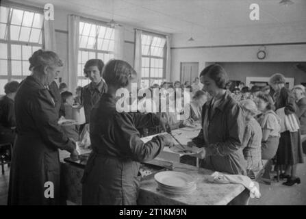 Il villaggio di invasione- La vita quotidiana in Orford, Suffolk, Inghilterra, 1941 membri delle donne del Volontariato (WVS) servire un pasto a persone presso il villaggio cucina comune, o ristorante britannico, in Orford. Dietro di loro, i bambini e gli adulti può essere visto a sistemare nella loro pranzo presso i lunghi tavoli in questo puramente funzionale sede. Foto Stock