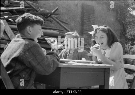 Aiuti dall America- Lend Lease cibo, Londra, Inghilterra, 1941 Un gruppo di bambini sorridenti sedersi a un tavolo e mangiare formaggio importato da America come parte del regime Lend-Lease nel parco giochi di una bomba gravemente danneggiati a scuola. La scuola è ora utilizzata come un centro di alimentazione. Pali di legno recuperato può essere visto solo dietro i bambini. Questa fotografia è stata probabilmente preso a fine agosto o primi di settembre 1941. Foto Stock