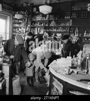 Onorevole Bugler va alla guerra - La vita quotidiana in Oriente Dean, Sussex, Inghilterra, 1943 Una vista interna Dennett, il villaggio occupato store in Oriente Dean. Onorevole Bugler può essere visto sulla destra della fotografia, guardare come un altro cliente gioca con un cane. Un altro cliente e il bambino anche guardare il cane come John vigilia, un evacuee, anelli fino al totale sul registro di cassa, supervisionato da Miss Absale (oscurata dalla onorevole Bugler's hat). Miss Absale è stato bombardato da Londra e ora lavora per tre giorni la settimana part-time nel villaggio, nonché in esecuzione il risparmio locale gruppo. L'uomo che lavora dietro il bancone (a destra di t Foto Stock