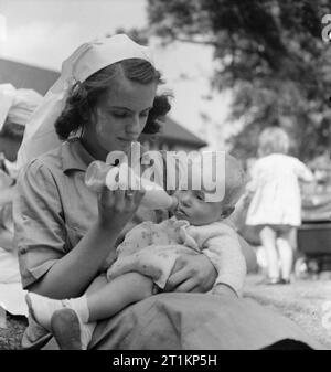 Nursery per le mamme che lavorano- il lavoro della Pietra Focaia Strada Verde vivaio, Birmingham, 1942 un giovane infermiere bottiglia alimenta un bambino in serata sole nel parco di pietra focaia Strada Verde vivaio. Foto Stock
