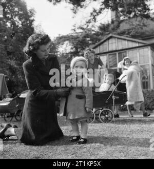 Nursery per le mamme che lavorano- il lavoro della Pietra Focaia Strada Verde vivaio, Birmingham, 1942 20 enne ra Ann Donovan serra i pulsanti sul suo figlio Michael del mantello e come essa raccoglie lui dal giardino del vivaio. Michael è di 16 mesi. Onorevole Donovan ha appena completato una giornata di lavoro in una fabbrica di munizioni. Dietro di loro un altro madre può essere visto la raccolta di sua figlia in una carrozzina e un infermiere porta un altro bambino a una diversa parte del giardino. Foto Stock