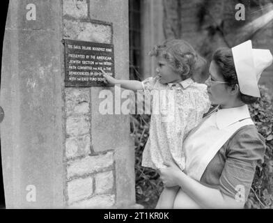 Gli aiuti americani per la Gran Bretagna- Vita a corte Syston nursery, Sara Delano Roosevelt Home, Gloucestershire, Inghilterra, 1942 Un infermiere tiene un bambino in modo che essa è in grado di leggere la targhetta del nome del vivaio a cui ella è il vostro soggiorno. La placca si legge: "Sara Delano Roosevelt Home. Presentato dalla American Jewish Sezione della commissione interreligiosa per gli aiuti a favore della democrazia in cooperazione con il Fondo di Esco comitato'. Foto Stock