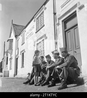 Le truppe USA in un Villaggio Inglese- La vita di tutti i giorni con gli americani in Burton Bradstock, Dorset, Regno Unito 1944 Quattro soldati americani sedervi al sole sul marciapiede fuori del Anchor Inn in Burton Bradstock e chat per ragazza locale Betty 'lentiggini' Mackay. Secondo la didascalia originale, il soprannome di "lentiggini' è stato dato a Betty da GIs, che hanno 'made di lei un accampamento favorito". Foto Stock