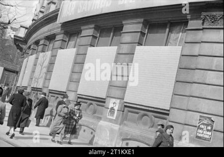 Blackout e precauzioni di raid aereo a Londra, Inghilterra, 1941 Le finestre di questo grande edificio arrotondato (probabilmente Westminster City Hall su Charing Cross Road) sono state murate, sia come un modo per blackout dell'edificio, ma anche per evitare che il vetro da essendo continuamente devastata da incursioni aeree. Vari poster, compreso un annuncio pubblicitario per la guerra di risparmio, può essere visto sulla costruzione e diversi membri del pubblico a piedi passato come andare circa le loro attività quotidiane. Foto Stock