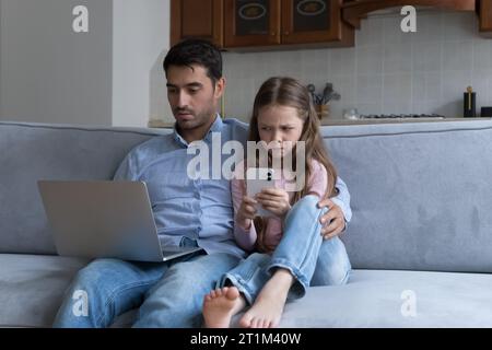 Papà addicted di gadget che usa il laptop a casa, abbracciando Foto Stock