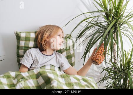 Un bambino di 9-10 anni, con i suoi capelli a cascata, si sdraia a letto, tenendo delicatamente un ananas, incarnando l'essenza del relax e del tempo libero Foto Stock