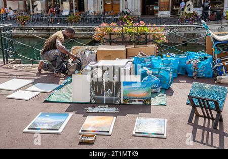 Un venditore di strada si prepara a realizzare e mostrare immagini dipinte a spruzzo in vendita come souvenir turistici su un marciapiede nel centro di Annecy, in Francia Foto Stock