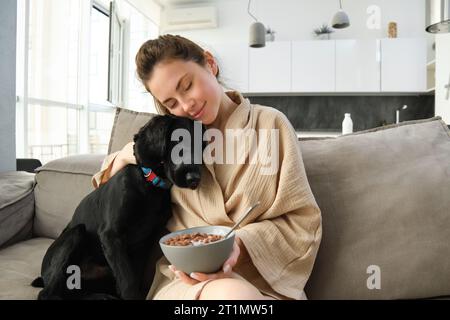 donna con i capelli corti seduta su un divano a casa, che mangia cereali a colazione e accarezza il suo amato cane. Il cane sta annusando una ciotola di cereali Foto Stock